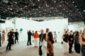 Visitors walk through a busy contemporary art fair, browsing paintings and sculptures displayed on white gallery walls
