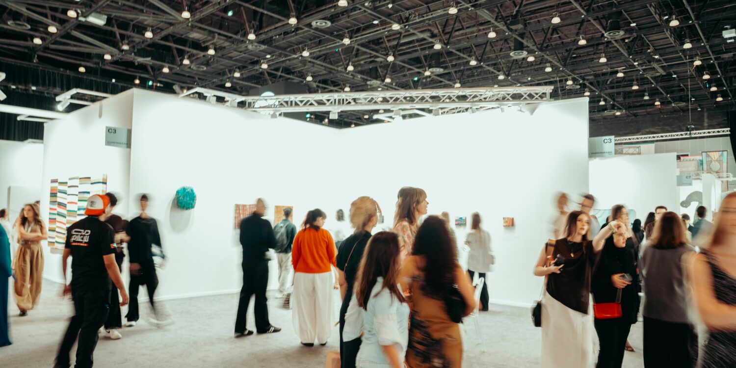 Visitors walk through a busy contemporary art fair, browsing paintings and sculptures displayed on white gallery walls