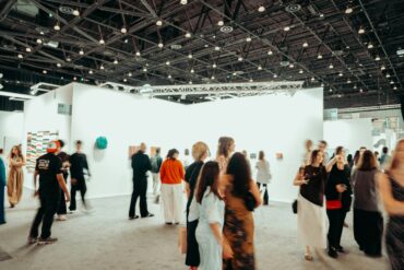 Visitors walk through a busy contemporary art fair, browsing paintings and sculptures displayed on white gallery walls