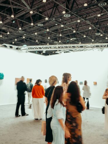 Visitors walk through a busy contemporary art fair, browsing paintings and sculptures displayed on white gallery walls