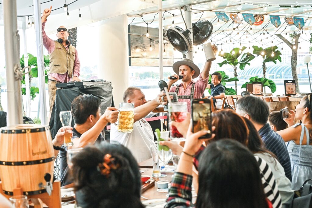 Live entertainment and crowd toast at Brotzeit Singapore's Oktoberfest celebration, with DJ and Bavarian-costumed performer on stage