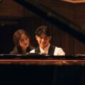 George Harliono and Hera Hyesang Park sit together at a grand piano, both looking down at the keys as he smiles