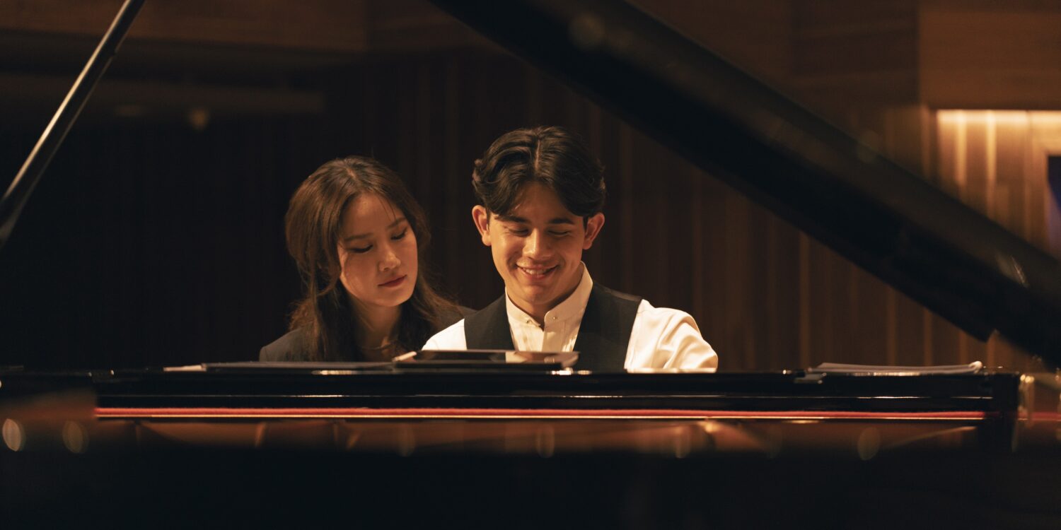 George Harliono and Hera Hyesang Park sit together at a grand piano, both looking down at the keys as he smiles