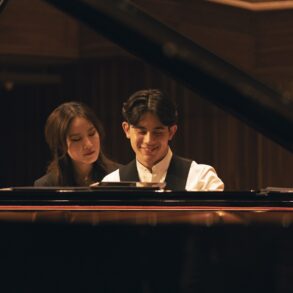George Harliono and Hera Hyesang Park sit together at a grand piano, both looking down at the keys as he smiles