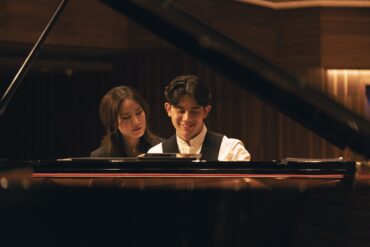 George Harliono and Hera Hyesang Park sit together at a grand piano, both looking down at the keys as he smiles