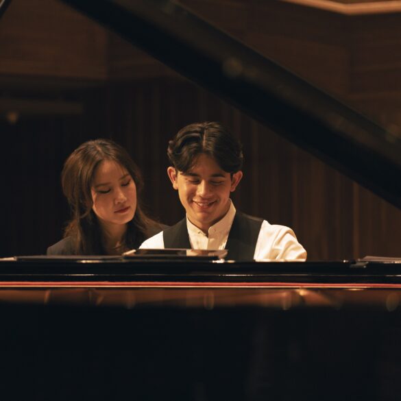 George Harliono and Hera Hyesang Park sit together at a grand piano, both looking down at the keys as he smiles