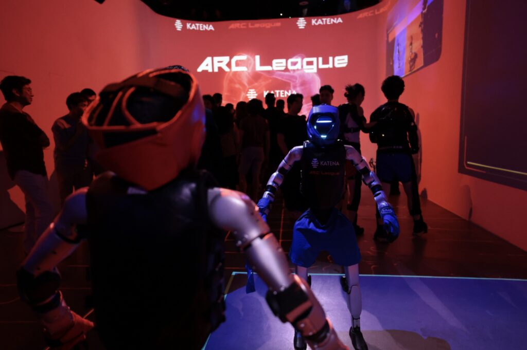 Two humanoid robots in boxing gear face off in an arena at an ARC League Robot Fight Night event, with a crowd gathered behind an ARC League screen