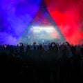 Silhouetted crowd dances beneath a triangular stage lit in blue and red smoke at a Rainbow Disco Club festival, with a DJ performing inside the illuminated booth.