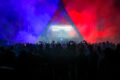 Silhouetted crowd dances beneath a triangular stage lit in blue and red smoke at a Rainbow Disco Club festival, with a DJ performing inside the illuminated booth.