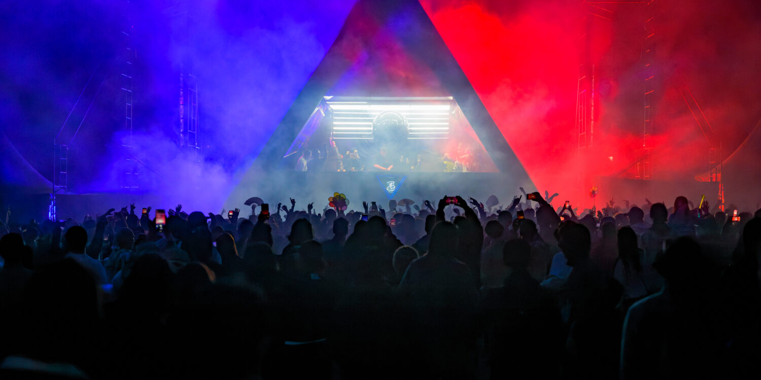 Silhouetted crowd dances beneath a triangular stage lit in blue and red smoke at a Rainbow Disco Club festival, with a DJ performing inside the illuminated booth.