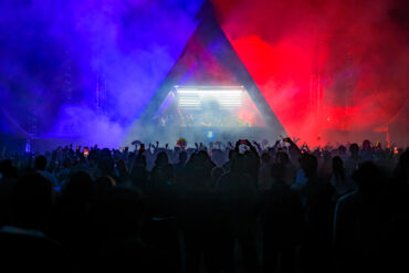 Silhouetted crowd dances beneath a triangular stage lit in blue and red smoke at a Rainbow Disco Club festival, with a DJ performing inside the illuminated booth.
