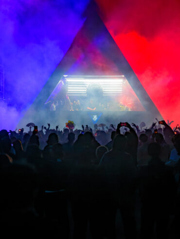 Silhouetted crowd dances beneath a triangular stage lit in blue and red smoke at a Rainbow Disco Club festival, with a DJ performing inside the illuminated booth.