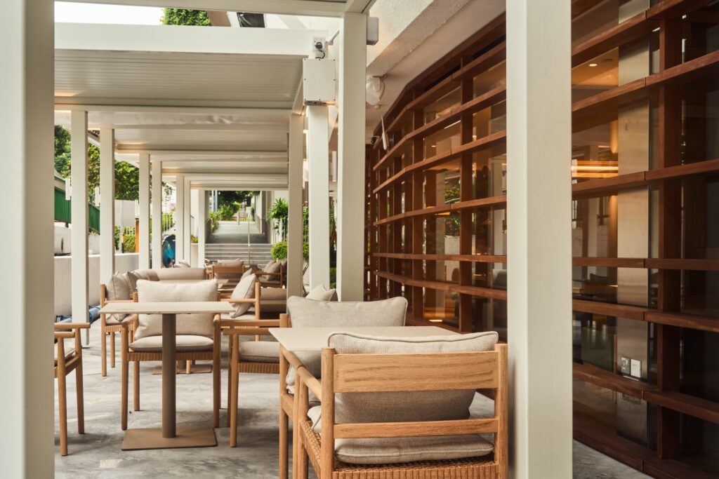 Al fresco terrace at AO, 1 Claymore Drive, with wooden armchairs, linen cushions and stone tables beside a timber-framed façade.