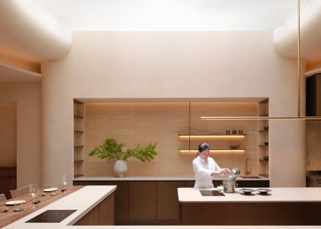 Chef Johanne Siy at the open kitchen counter of Árō on Mohamed Sultan Road, under a skylight with woven mesh and travertine shelving.