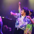 Performers dance on stage under purple lights at DARE Festival, with a performer in glasses and a blue floral shirt in front and dancers in kebaya behind.