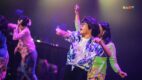 Performers dance on stage under purple lights at DARE Festival, with a performer in glasses and a blue floral shirt in front and dancers in kebaya behind.