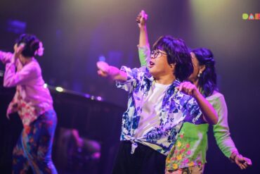 Performers dance on stage under purple lights at DARE Festival, with a performer in glasses and a blue floral shirt in front and dancers in kebaya behind.