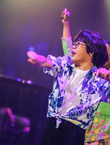 Performers dance on stage under purple lights at DARE Festival, with a performer in glasses and a blue floral shirt in front and dancers in kebaya behind.