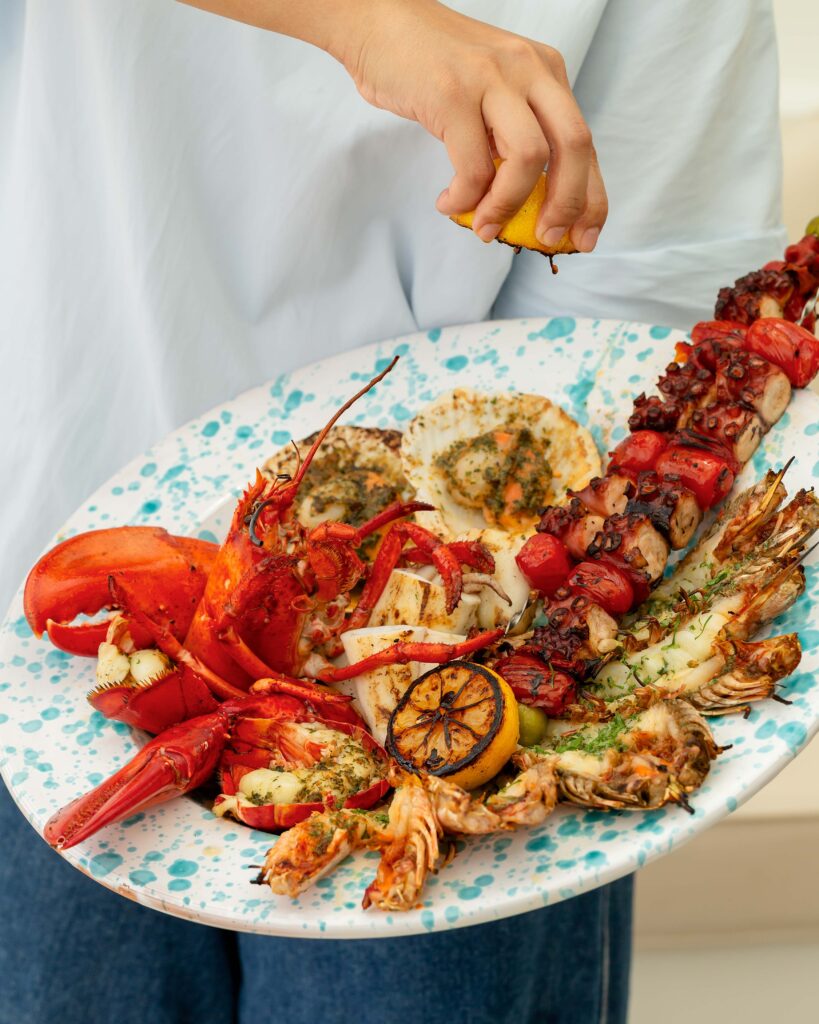 Charcoal-grilled seafood platter at Fico, Tanjong Beach Club, with lobster, scallops, prawns and an octopus skewer on a speckled plate.