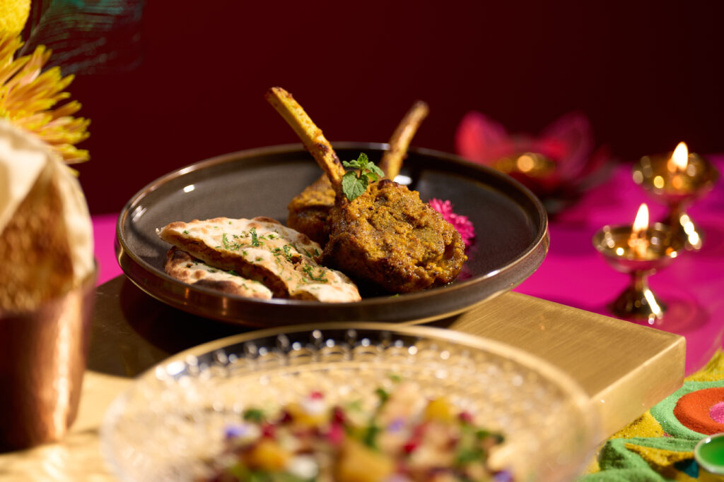 Achari Tandoori Lamb chops with keema kulcha from Tiffin Room’s Deepavali menu at Raffles Hotel Singapore, with brass diyas and a rangoli behind.