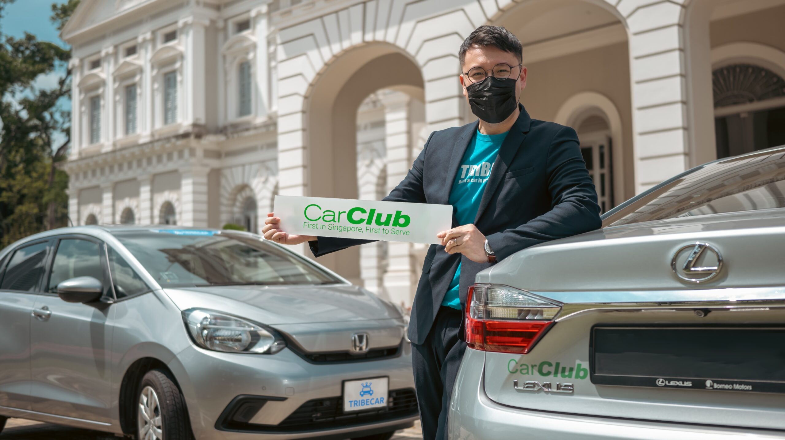 Tribecar co-founder Adrian Lee holding a Car Club sign while leaning on a silver Car Club Lexus, beside a Tribecar Honda, outside a colonial-era building in Singapore.
