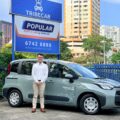 Tribecar co-founder Adrian Lee, standing beside a grey-green Tribecar car sharing MPV, in front of Tribecar and Popular Rent A Car by Tribecar signage in Singapore.