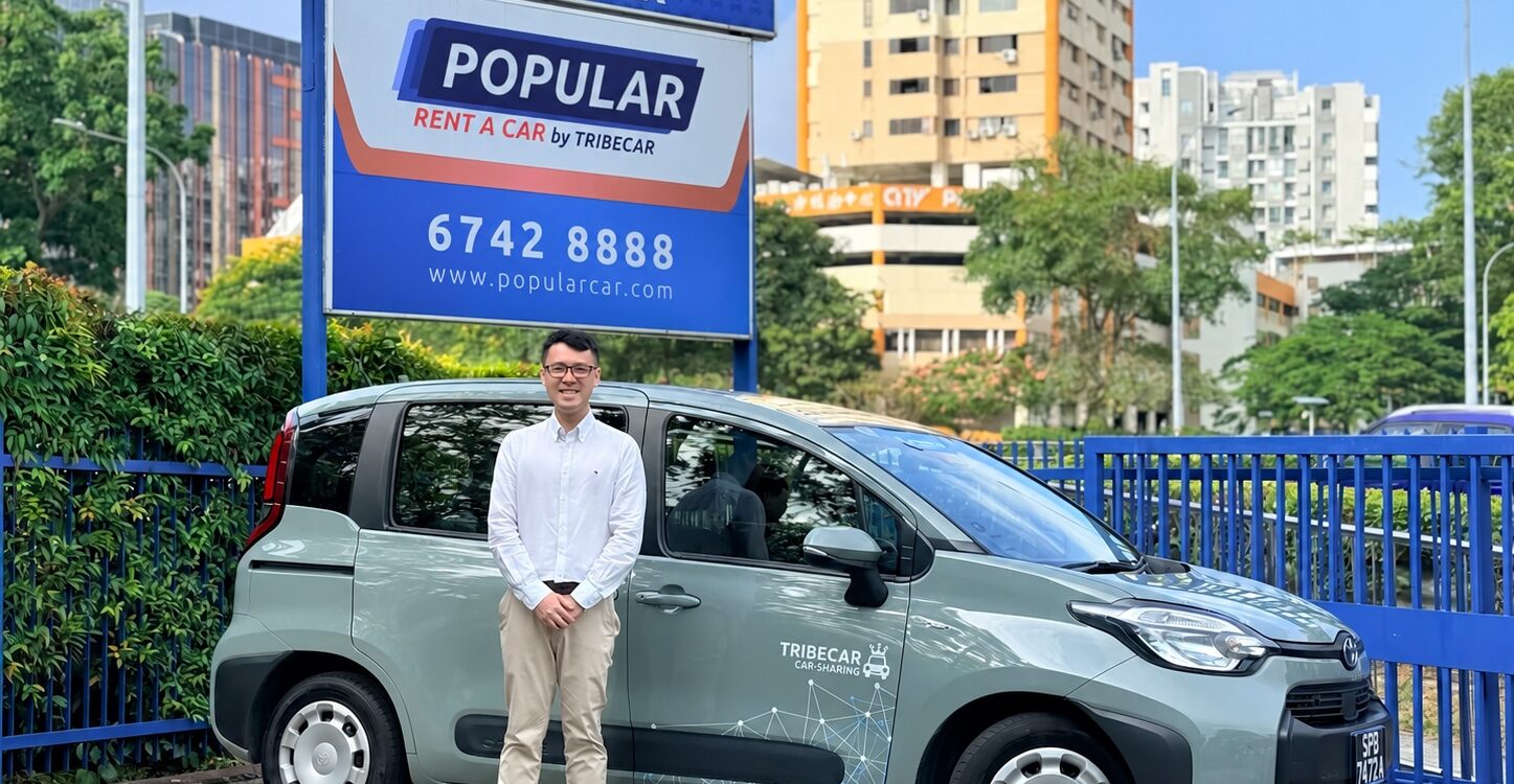 Tribecar co-founder Adrian Lee, standing beside a grey-green Tribecar car sharing MPV, in front of Tribecar and Popular Rent A Car by Tribecar signage in Singapore.