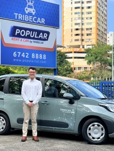 Tribecar co-founder Adrian Lee, standing beside a grey-green Tribecar car sharing MPV, in front of Tribecar and Popular Rent A Car by Tribecar signage in Singapore.