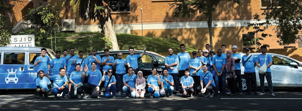 Tribecar volunteers in blue Tribecar T-shirts posing with Tribecar vehicles outside a mosque in Singapore during a Ramadan volunteer drive.