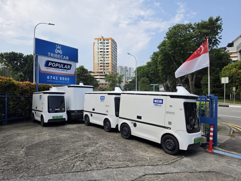 Four Neolix autonomous delivery vehicles parked outside Tribecar and Popular Rent A Car by Tribecar signage in Singapore, beside a Singapore flag.