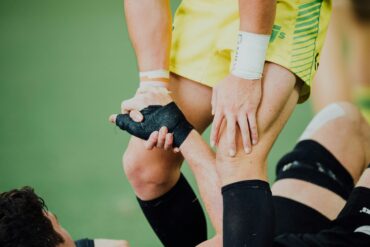 Rugby player in yellow shorts pulling a fallen opponent up by his taped hand on a grass pitch