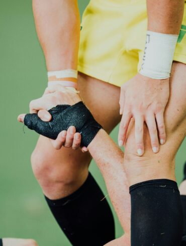 Rugby player in yellow shorts pulling a fallen opponent up by his taped hand on a grass pitch