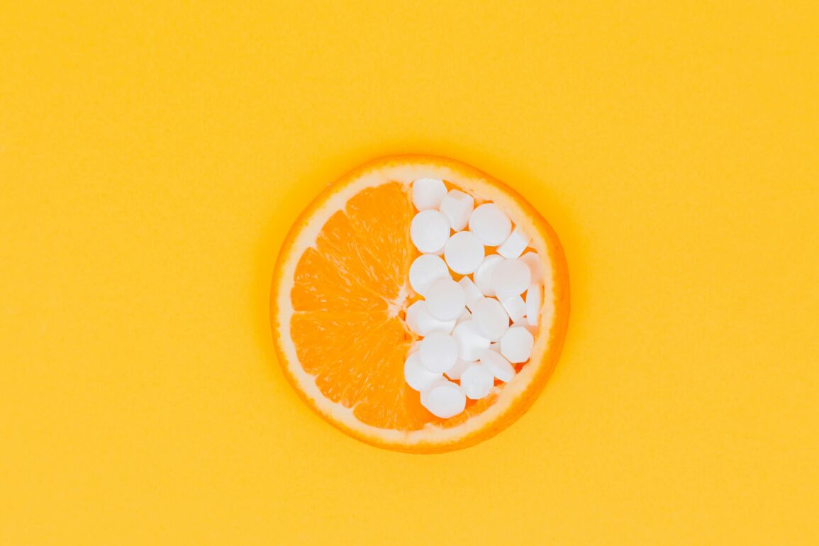 Orange slice with half its flesh replaced by white supplement tablets on a yellow background.