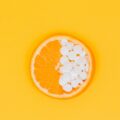 Orange slice with half its flesh replaced by white supplement tablets on a yellow background.