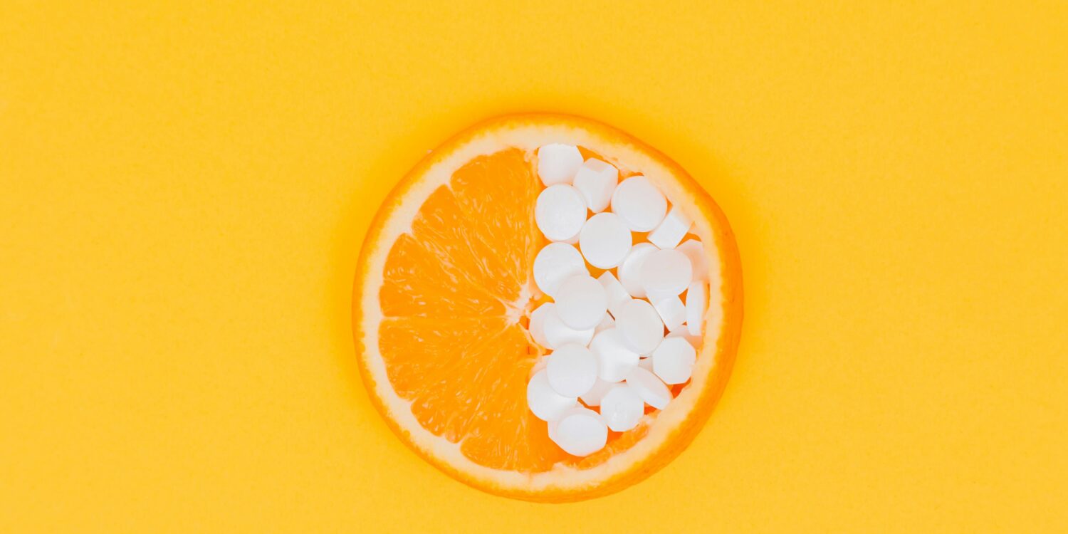 Orange slice with half its flesh replaced by white supplement tablets on a yellow background.