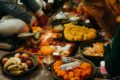 A family sits on the floor around a lit diya during a Deepavali prayer, surrounded by marigold garlands on leaf plates, a box of mithai, brass pots and offerings of fruit.