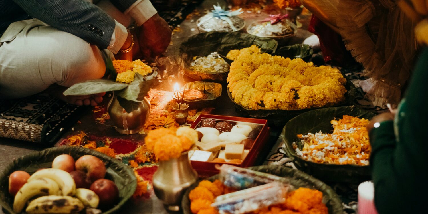A family sits on the floor around a lit diya during a Deepavali prayer, surrounded by marigold garlands on leaf plates, a box of mithai, brass pots and offerings of fruit.