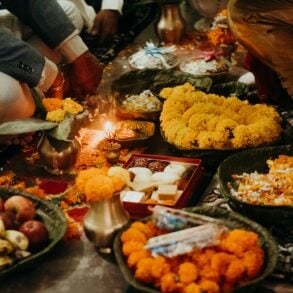 A family sits on the floor around a lit diya during a Deepavali prayer, surrounded by marigold garlands on leaf plates, a box of mithai, brass pots and offerings of fruit.