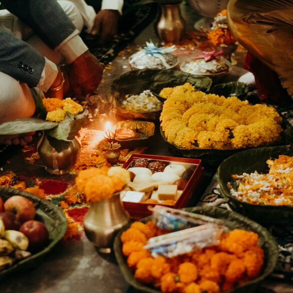 A family sits on the floor around a lit diya during a Deepavali prayer, surrounded by marigold garlands on leaf plates, a box of mithai, brass pots and offerings of fruit.